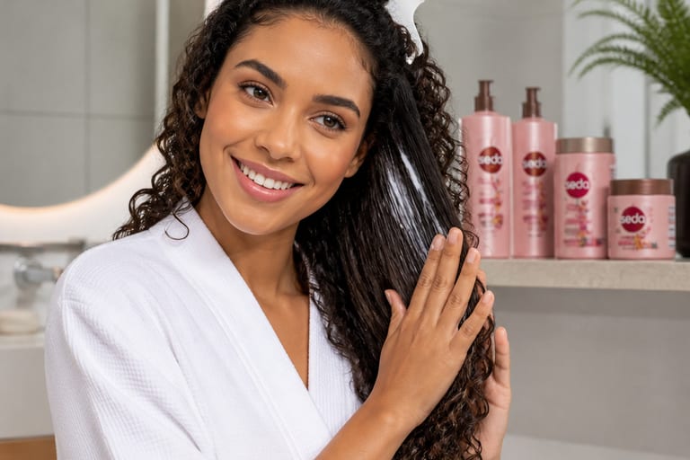 Mulher sorrindo enquanto aplica creme de pentear em cabelo cacheado em um banheiro bem iluminado, com produtos de cabelo organizados ao fundo.