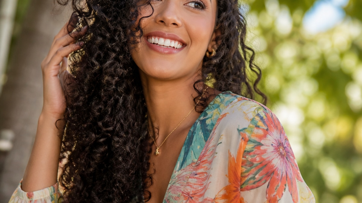 Mulher sorrindo com cabelo longo cacheado, vestindo blusa leve e colorida, em ambiente natural iluminado com árvores ao fundo.