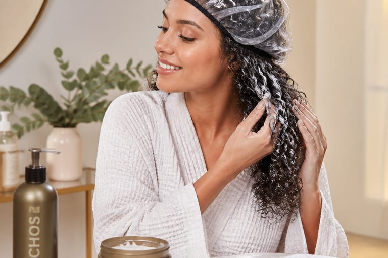 Mulher aplicando máscara capilar em cabelos cacheados, usando touca de hidratação, com produtos de cabelo ao redor, em ambiente bem iluminado.