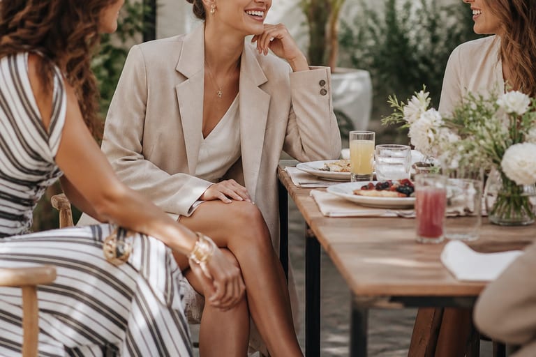 Mulher sorrindo e interagindo com amigas em um brunch ao ar livre, usando blazer oversize, vestido leve e sandálias em ambiente iluminado.