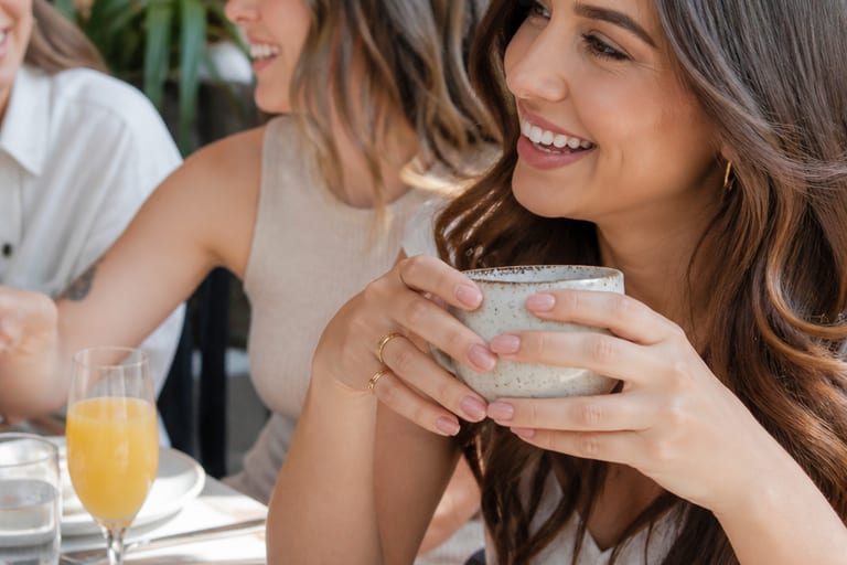 Mulher em brunch com amigas, mostrando unhas pintadas de esmalte nude em tom rosa queimado, em ambiente descontraído.
