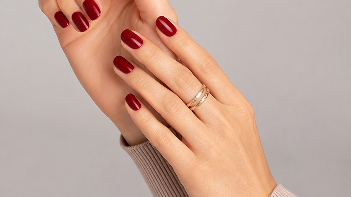 Mãos com unhas bem cuidadas e esmaltação vermelha escura em ambiente iluminado, com fundo neutro.