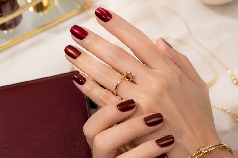 Mãos com esmalte vermelho escuro em acabamentos cremoso e metálico, com acessórios de moda ao fundo.