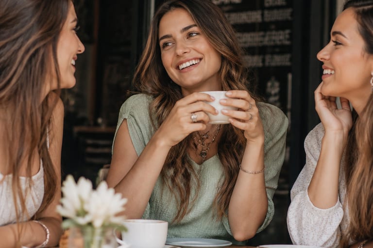 Mulher com maquiagem leve e natural, sorrindo e interagindo com amigas em um café.