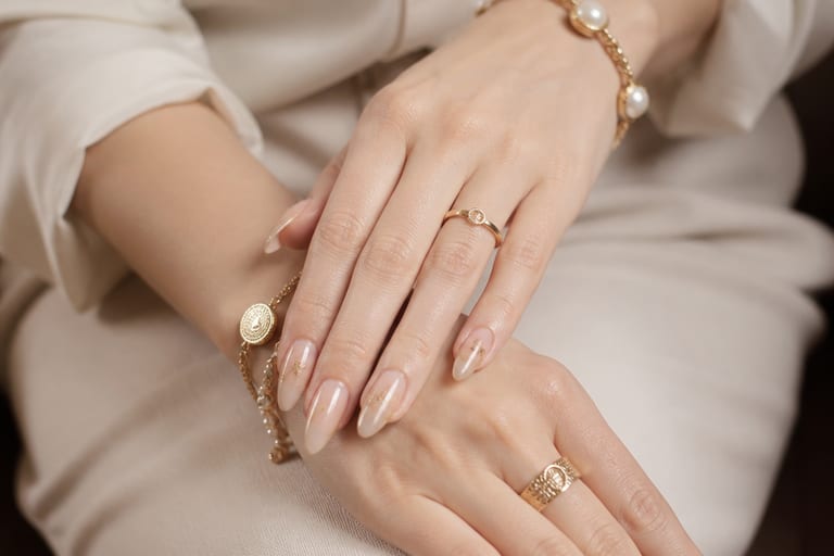 Mãos com unhas em gel decoradas, usando anéis e pulseiras, em um ambiente iluminado e elegante.