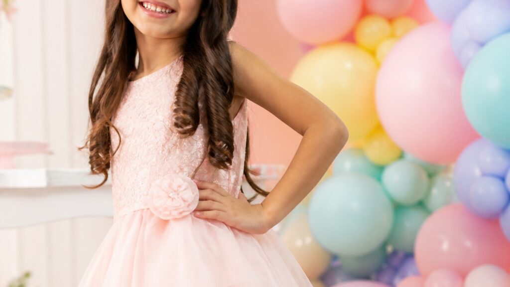 Menina sorridente usando um vestido infantil festa rosa claro em um ambiente iluminado com balões coloridos ao fundo.