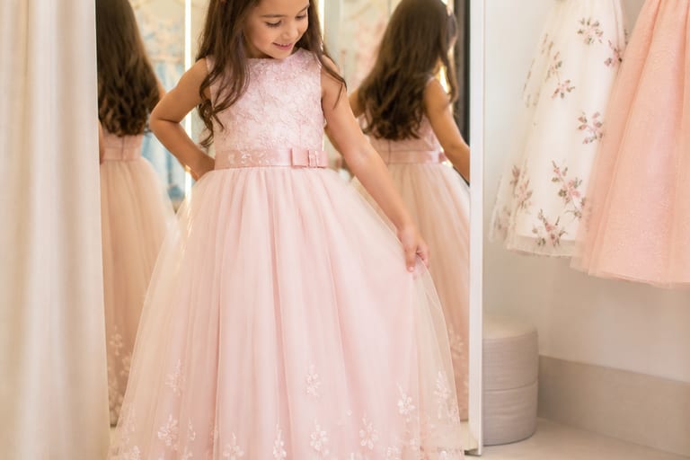 Menina experimentando vestidos infantis de festa em um provador, com espelhos ao fundo, em um ambiente iluminado e alegre.
