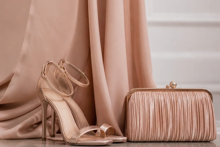 Close em sapatos de salto e clutch elegante ao lado de um vestido longo, em ambiente bem iluminado.